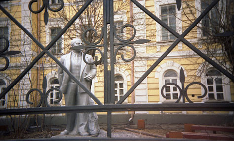 Looking through an ornate iron fence you see a grey statue of a man standing wearing a suit.  There is a yellow building in the background