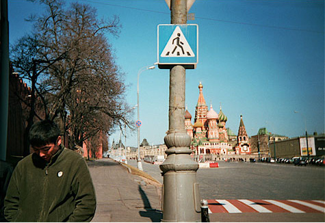 To the side, a man stands with his hands in his pockets, looking down.  Behind him is a street with a cross walk.  Farther back is an ornate building with colorful onion domes.