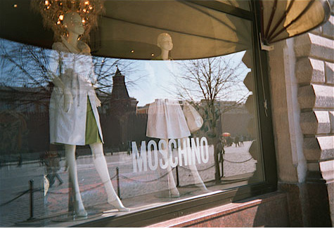Store window with two female mannequin's wearing dresses.  There is a reflection on the window of the street and an older building with a steeple