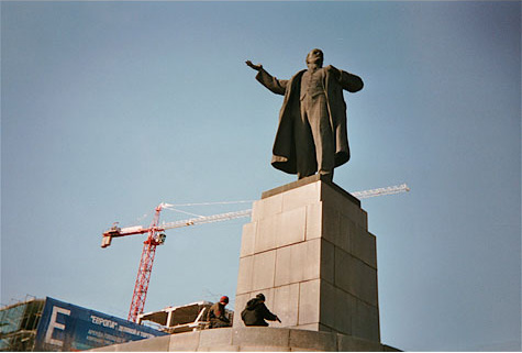 Looking up at men working at the base of a large monument of a man.  There is a large orange crane in the background and blue sky