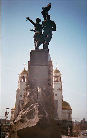 Looking up at a monument with a dramatic statue of a woman and man at the top.  There is a church in the background.