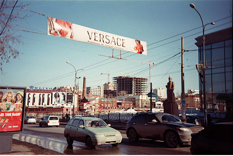 City scene with a statue of a man facing a busy street and advertisements on a banner and billboards