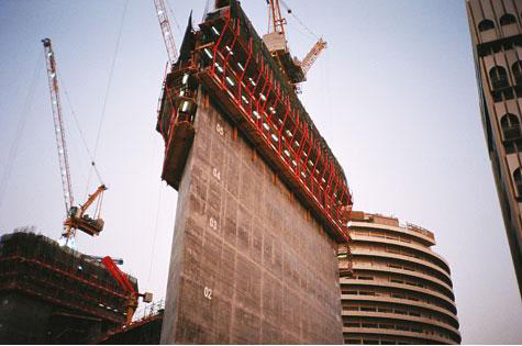 Photo looking up at a tall structure with cranes on top that is being built in the middle of a city