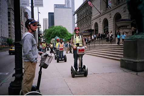 City scene with people standing on steps of a building and people heading down the sidewalk on two wheel balancing scooters.  One person in the forefront is stand to the side with their scooter