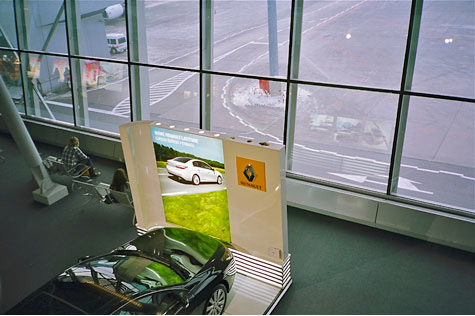 Interior of airport from above.  You see large windows that look onto the tarmac, a person waiting in a chair and a car on display