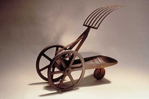 Metal sculpture of a cart with a fork like object protruding upwards by Bella Feldman