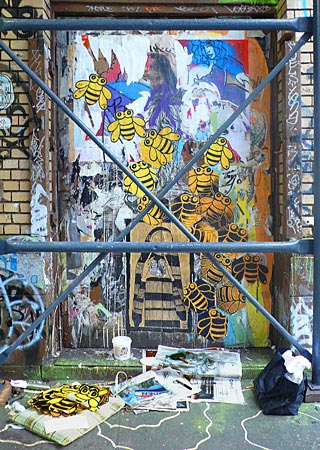 Image of abstract Street Art on door of a brick building.  Scaffolding stands in front of the door and art supplies are scattered on the ground.